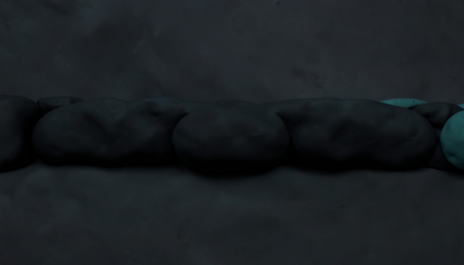 Dark, moody horizontal band of soft clay-model shapes in deep charcoal and muted teal, evoking an abstract studio scene at dusk.
